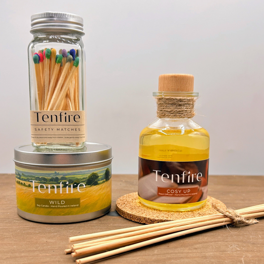 a glass jar of matches sitting on top of a candle tin and next to a glass bottle reed diffuser with rattan reeds in the foreground